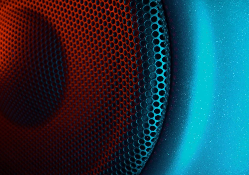Macro close-up of a professional studio monitor speaker showing the textured metal mesh grille and curved speaker cone under dramatic blue and orange lighting in a music production environment.