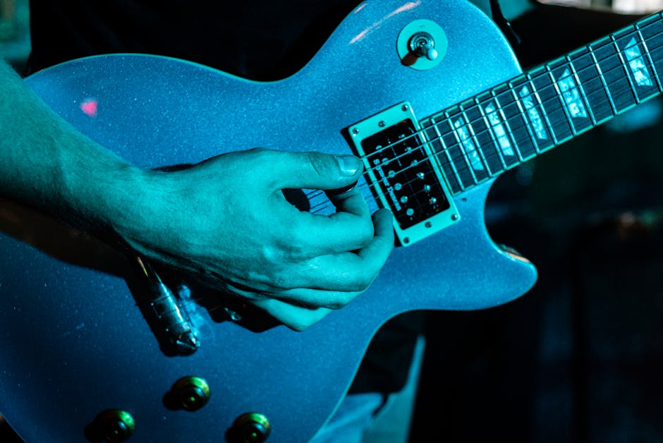 Close-up of a musician playing an electric guitar under blue stage lighting, focusing on the guitarist’s hand picking the strings near the pickups during a live music performance.